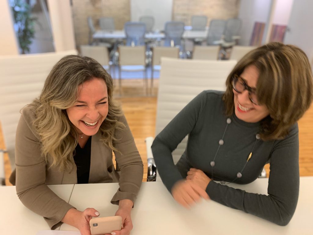 Two females sitting beside each other on white leather boardroom chairs, laughing, blurred action shot, with blurry background showing office space with another boardroom