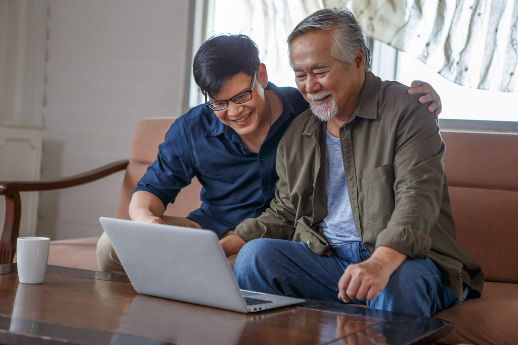 happy asian adult son and senior father sitting on sofa using laptop together at home . young man teaching old dad using internet online with computer on couch in living room .
