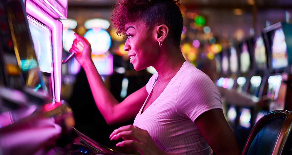woman from the side in a casino, pulling the handle on a slot machine with purple lights shining on her, so she looks purple