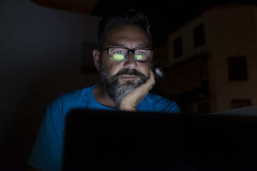 Stressed and worried man work by night on laptop computer - online social media life and stress for job people - adult male with glasses working with technology at home or office