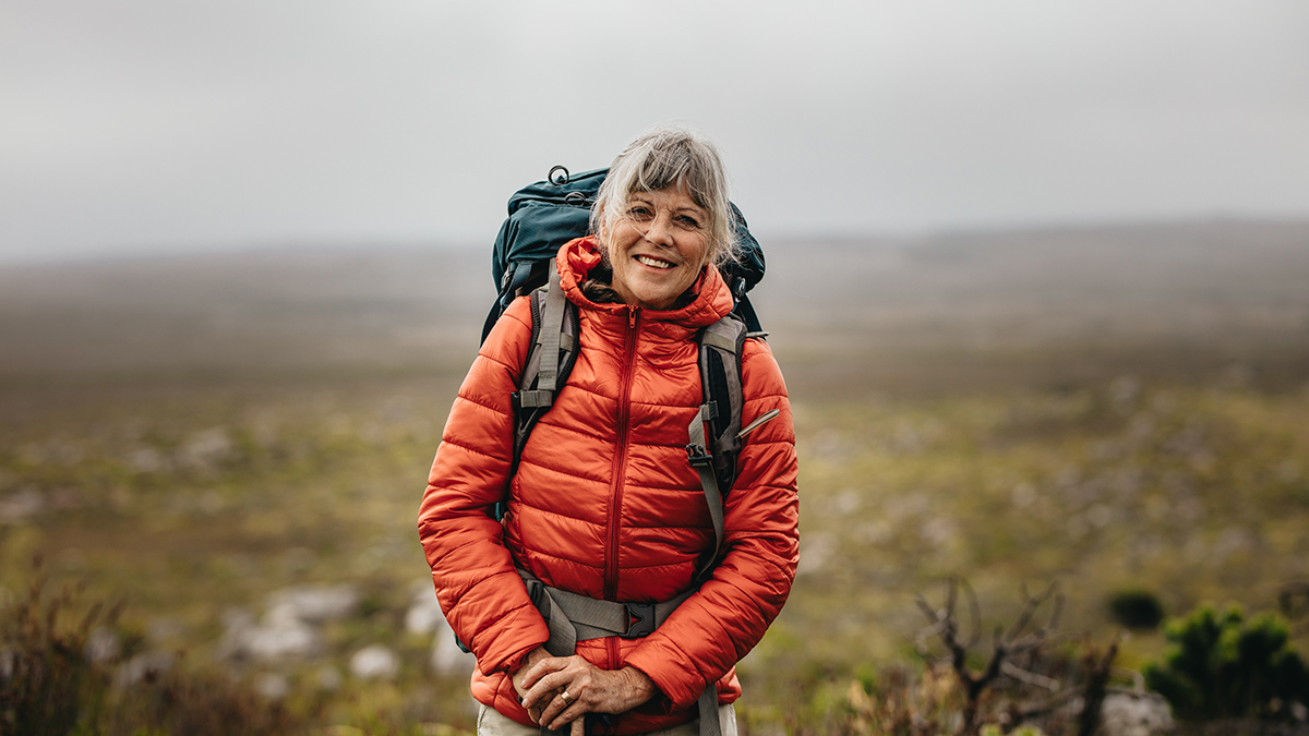 Portrait of a smiling female hiker standing on top of a hill on a winter day. Senior woman wearing jacket and backpack hiking on a hill.