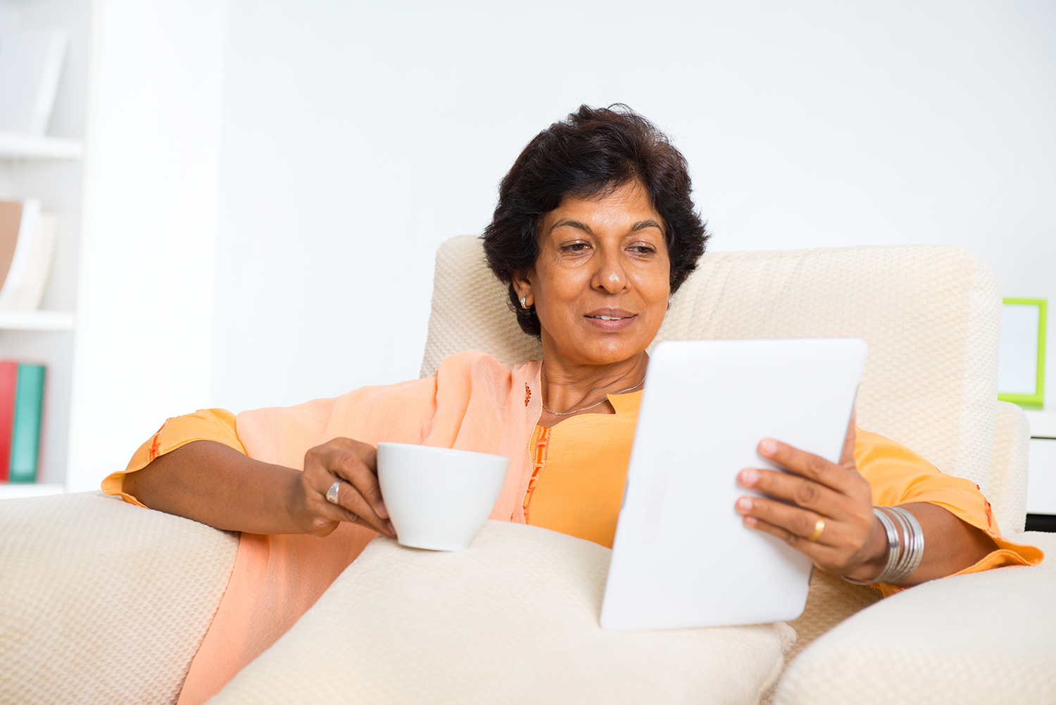 Mature 50s Indian woman drinking coffee / tea and using digital computer tablet at home