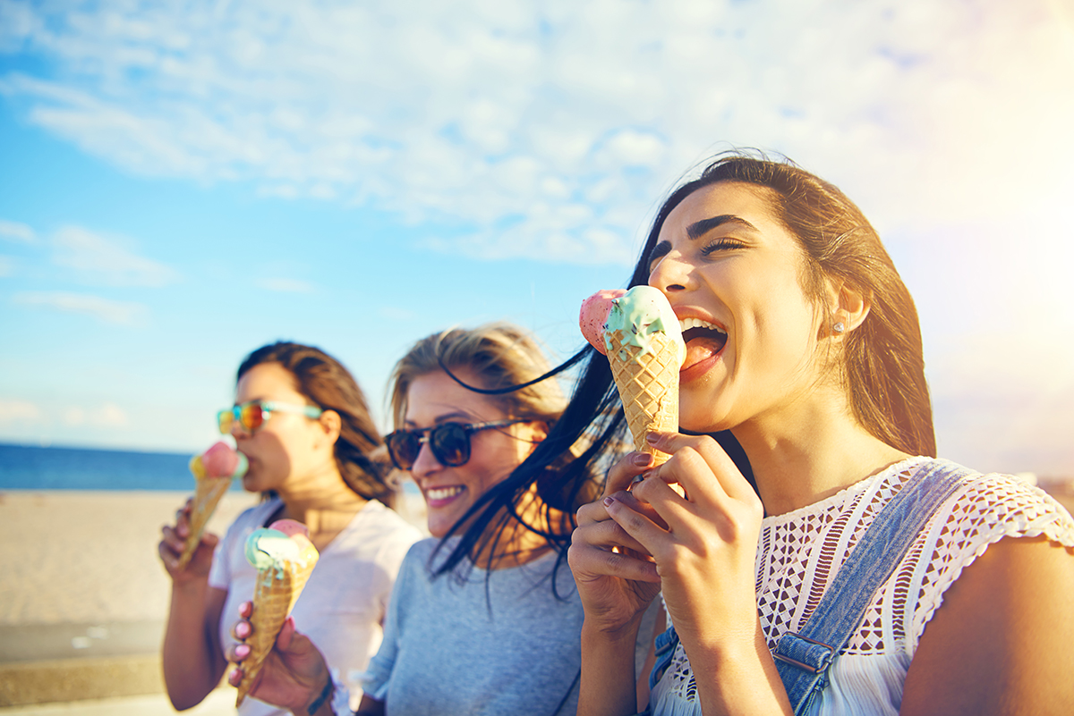three women eathing brightly coloured ice cream in cones while walking near ocean, sunny blue skies