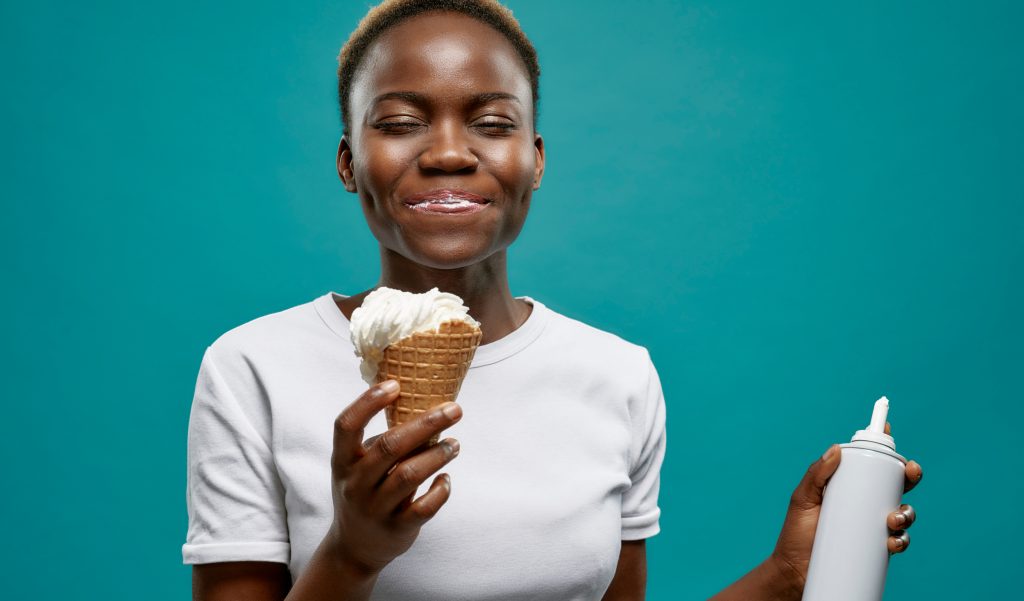 female with short hair, wearing a white t-shirt, standing in front of a green background, enjoying an ice cream in waffle cone, while holding whipped cream canister