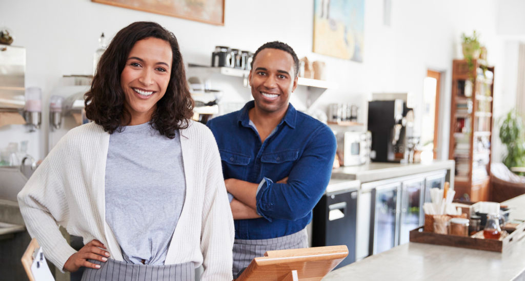 male and female cafe or coffee shop owners, wearing aprons, smiling while standing behind counter, light-filled modern cafe