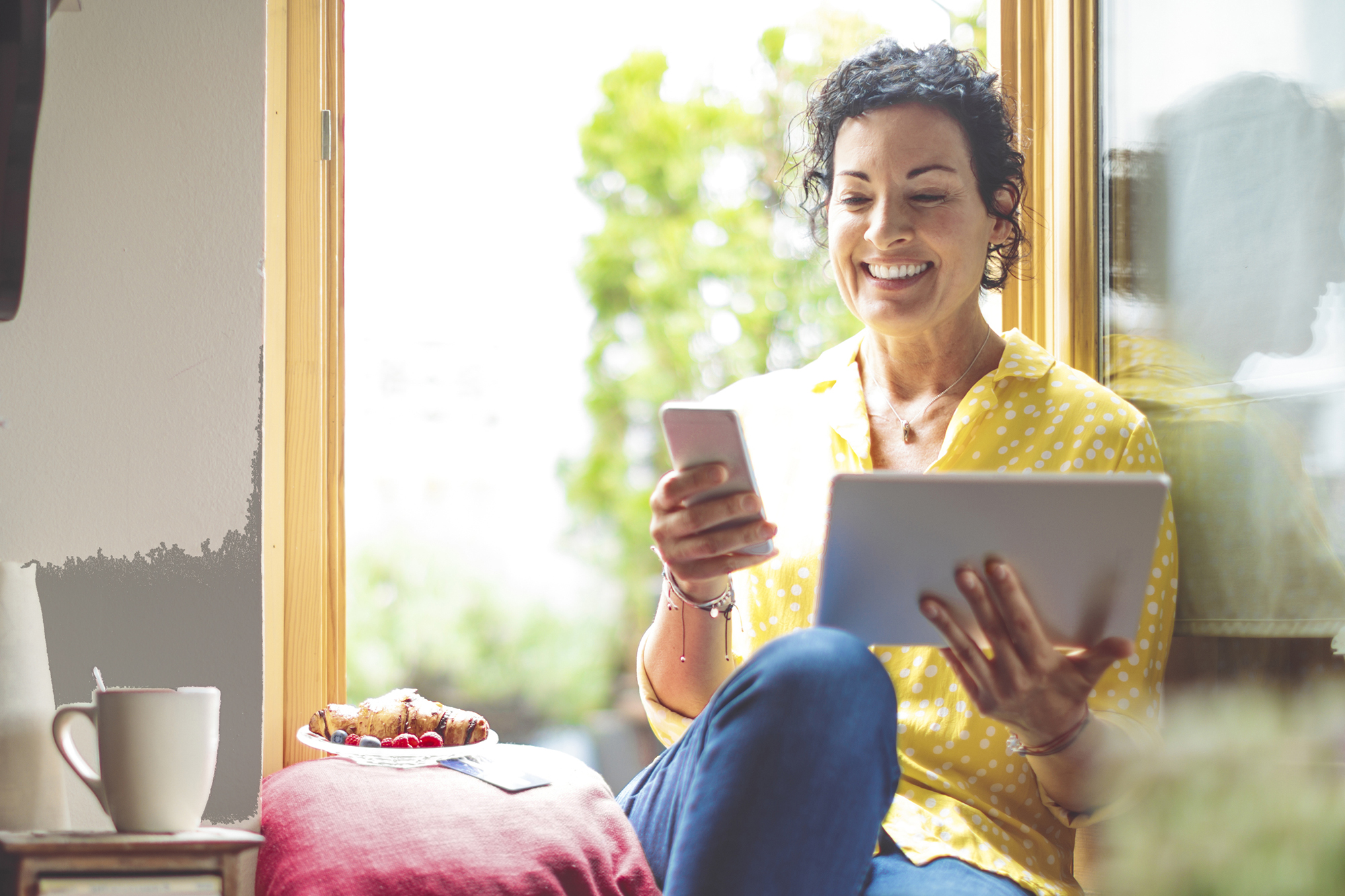 Mature woman is purchasing online at home - using mobile phone and tablet - while sitting in yellow window, wearing yellow shirt, with a plate of pastry and coffee mug