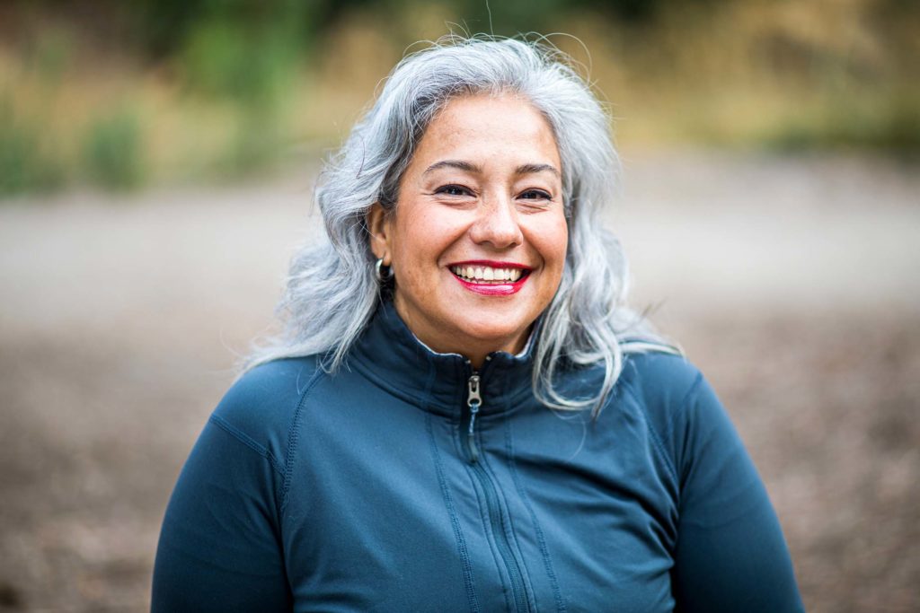close up of female with grey hair wearing blue zip-up excersie jacket, smiling, against a blurry background of nature