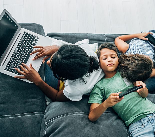 Mixed race family sitting on the sofa at home and using digital devices. African-american mother using laptop while her two children using smartphones.