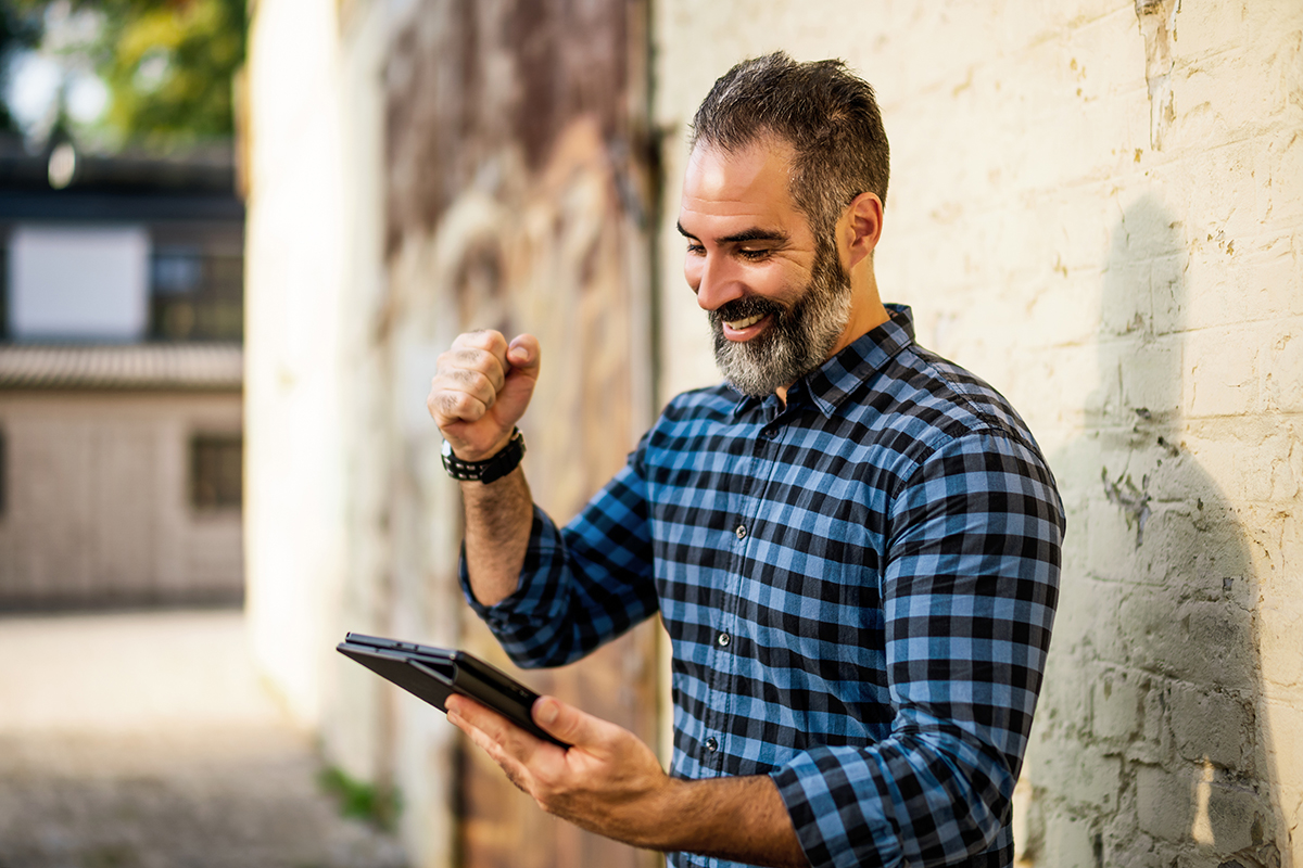 Excited modern businessman with beard using digital tablet while standing in front of brick wall outdoor.