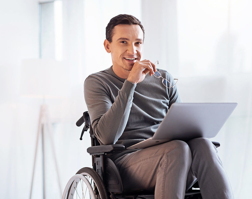 Having secret. Enigmatical male keeping smile on his face and holding glasses in right hand while sitting on the wheelchair