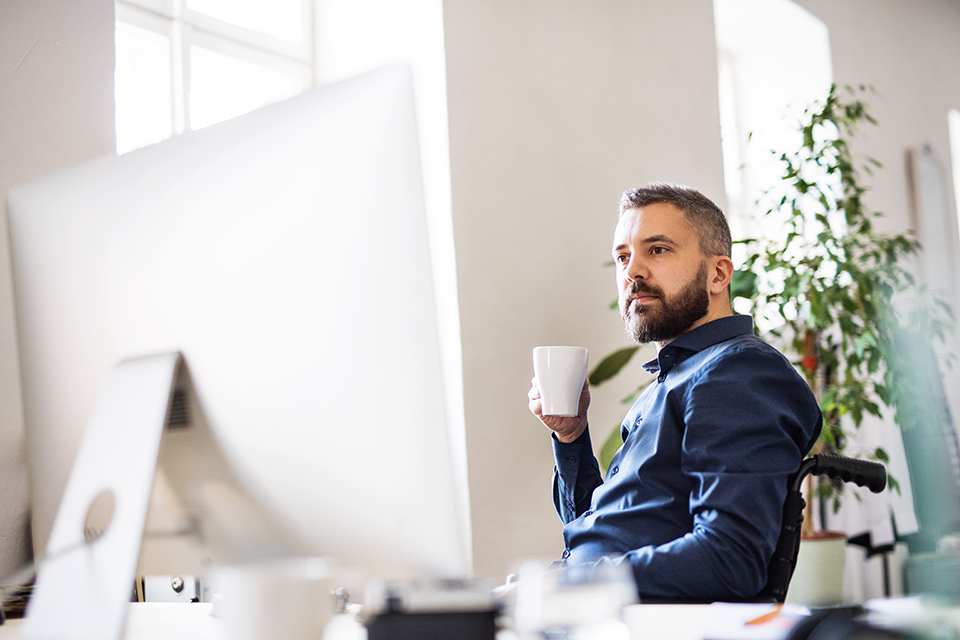 male sitting in wheelchair at desk, holding a coffee mug, wearing ablue shirt, while reading a computer monitor, light-filled modern office