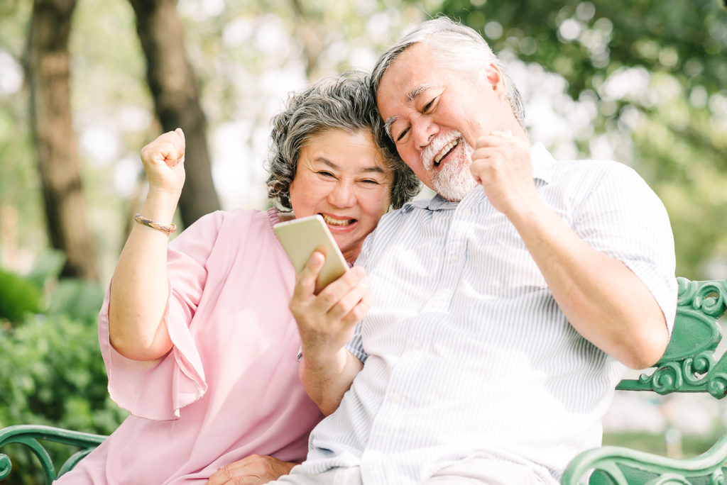 Happy senior Asian couple laughing and celebrating success together with smartphon in park