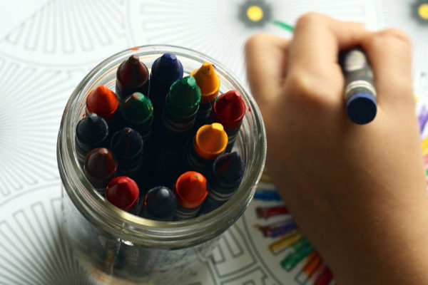 child's hand holding crayon, colour, with a jar full of crayons beside hand