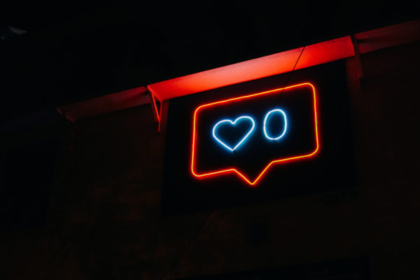 neon sign hanging off of building, at night, scene pitch black, neon sign is in a speech bubble in red with heart and number 0 in blue
