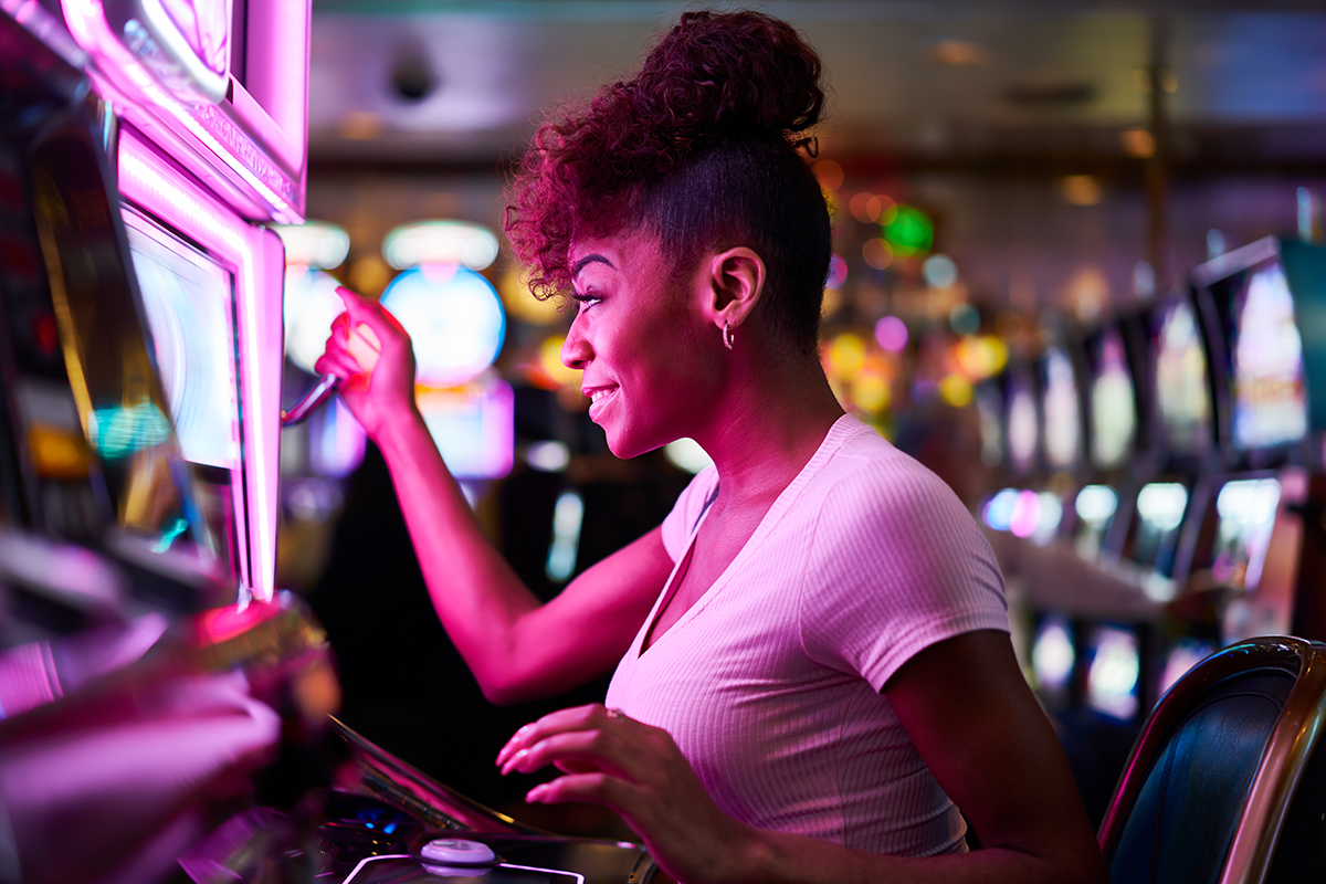 Woman from the side, sitting in a casino, with purple lights on her so she looks purple, pulling handle of slot machine