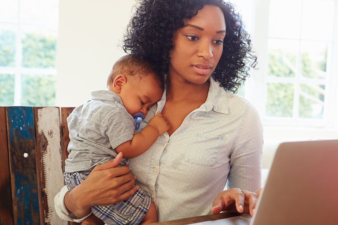 Mother holding infant against chest while using laptop on table, lots of light coming in windows