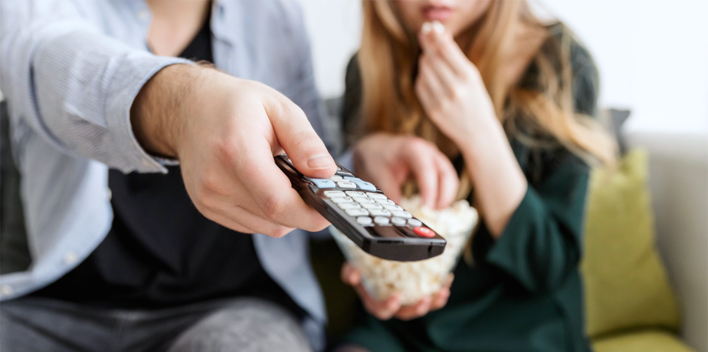 close up of male and female on couch eating popcorn and using the remote to change channels on TV