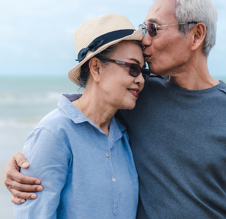 Senior couple, male with his arms wrapped around female, comforting her, both standing on a beach with blurry background
