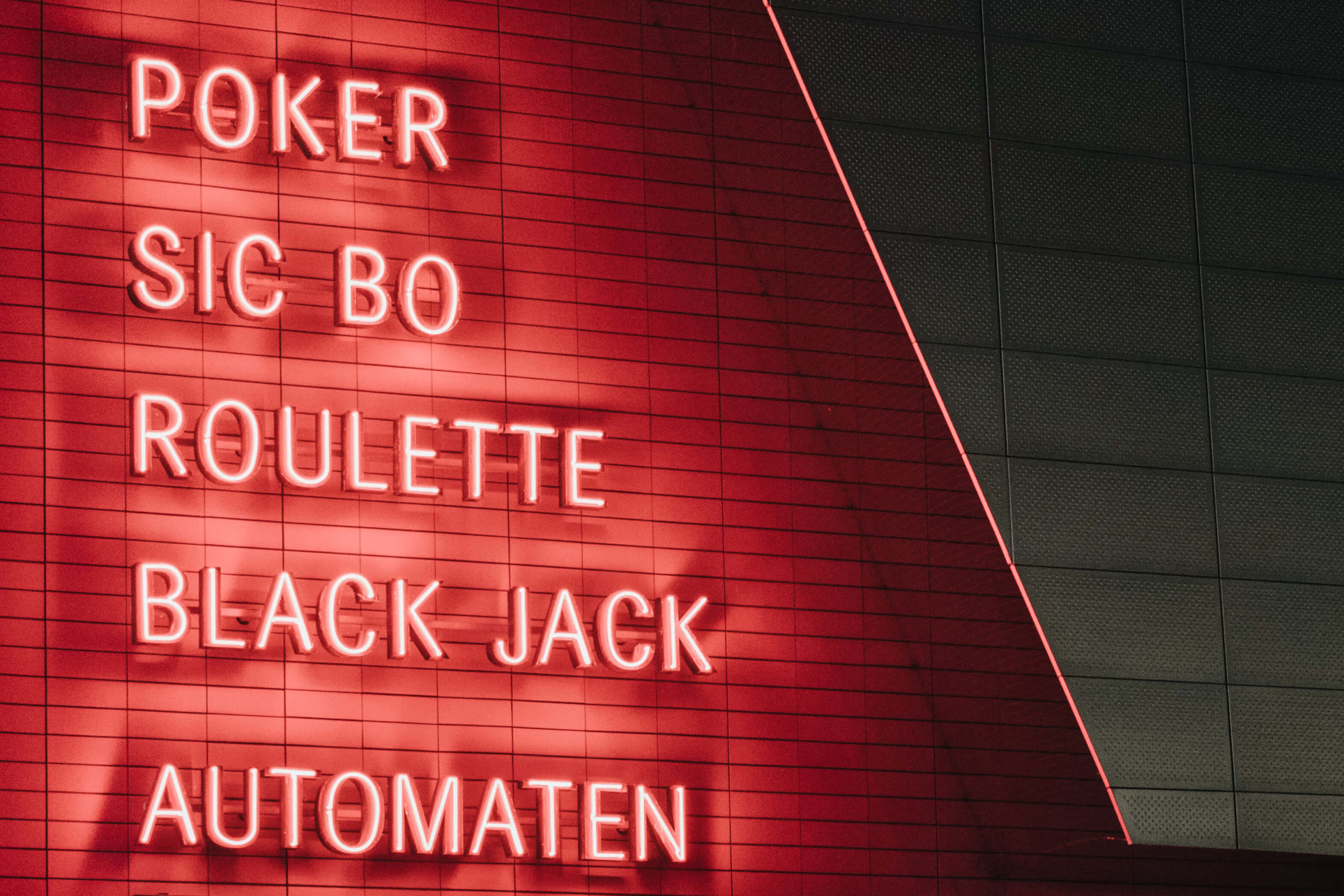 neon sign with red lettering on side of building with words: poker, sic bo, roulette, black jack, automaten